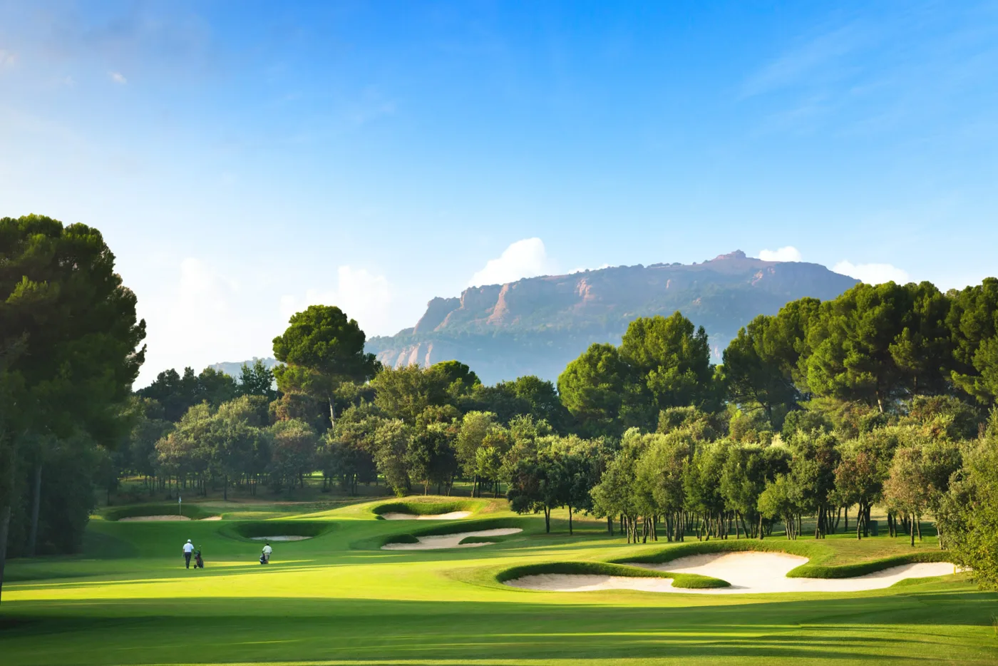 A pine-lined fairway and bunkered green at Real Club de Golf El Prat, with the mountains above Terrassa behind